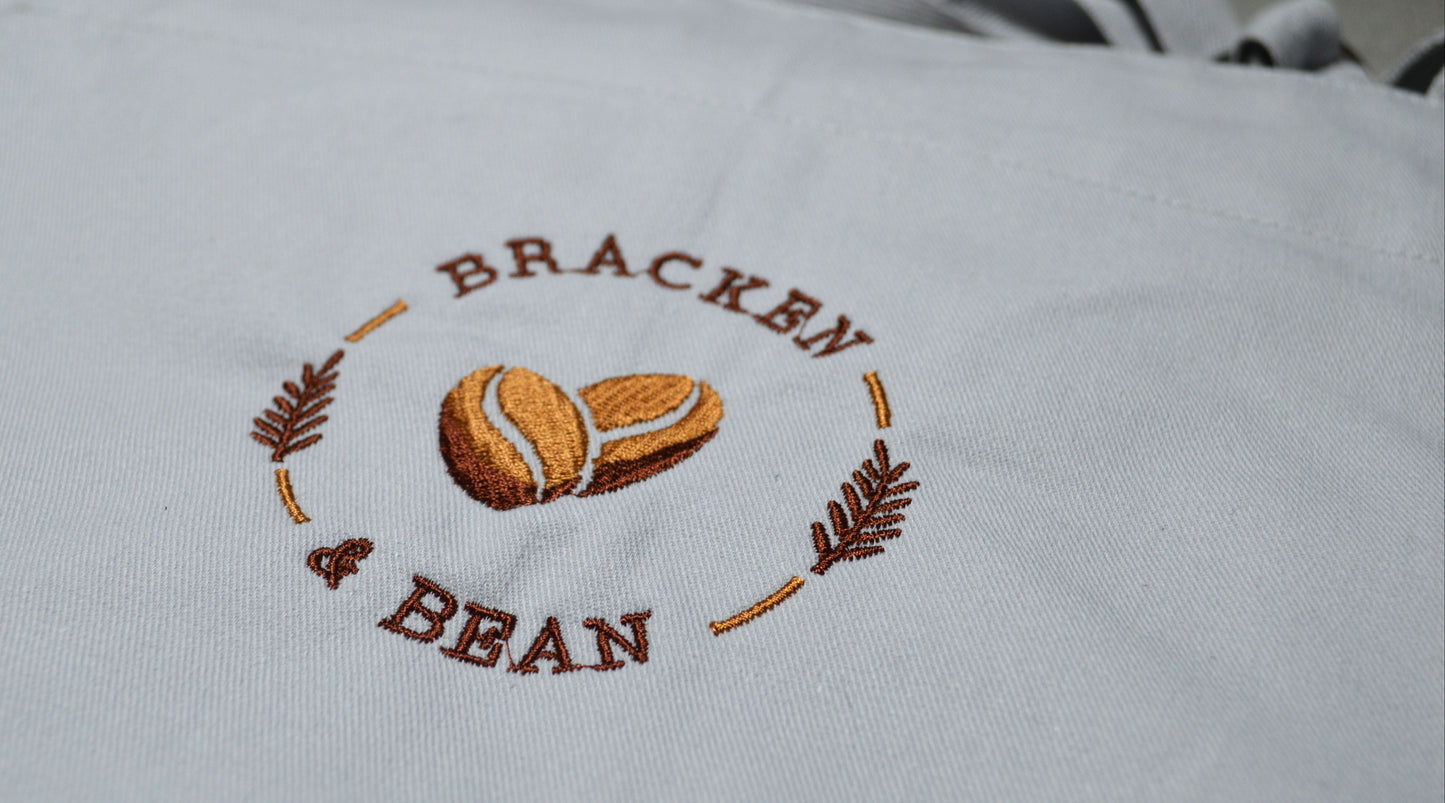 White apron with embroidered 'Bracken & Bean' logo on a white background